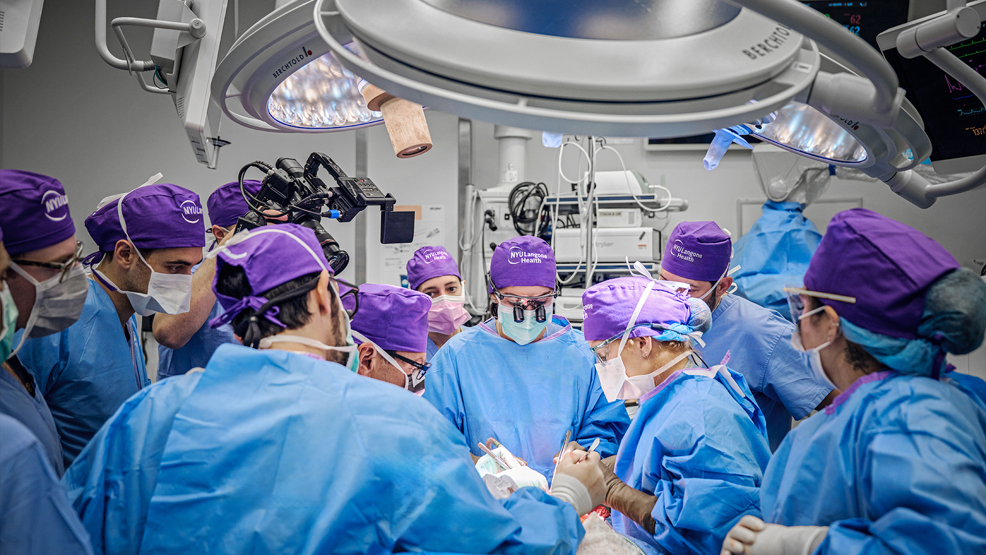 Doctors in blue medical gowns and purple surgical hats perform a surgery in an operating room.