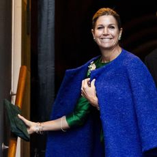 Queen Maxima wearing a blue coat holding a green clutch and smiling