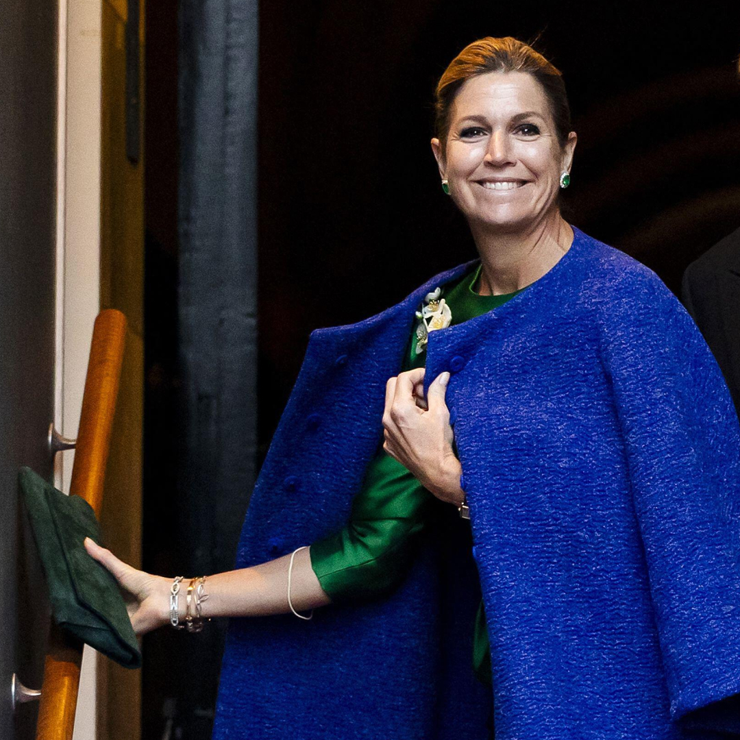 Queen Maxima wearing a blue coat holding a green clutch and smiling