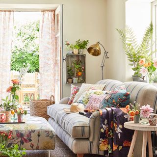 neutral living room with floral accents such as floral footstool, cushions and throw, a blue and cream striped sofa, and flowers and pot plants around the room