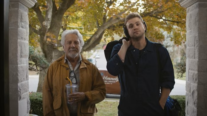 Two men stand outside a door frame. On the left is Niki, who has sandy brown hair and wears a black boiler suit. On the right is Harry, an older man with grey hair.