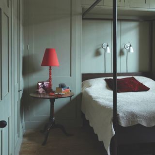 green grey bedroom with painted cabinetry and skirtings in the same shade, vintage side table, red lamp, four poster bed, cream bedspread, white wall lights