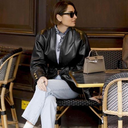 a French woman sits at an outdoor cafe in Paris wearing a leather jacket, neck scarf, and jeans