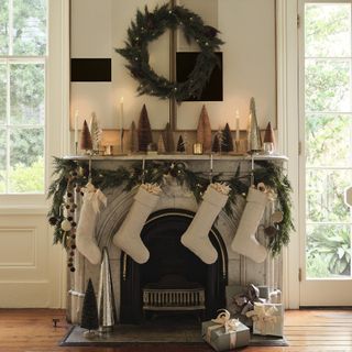 White living room with a marble and black fireplace. The mantel is decorated with neutral and classic Christmas decor. There is garland, four off-white stockings, a wreath above it, and multicolored decorative trees on the mantel.