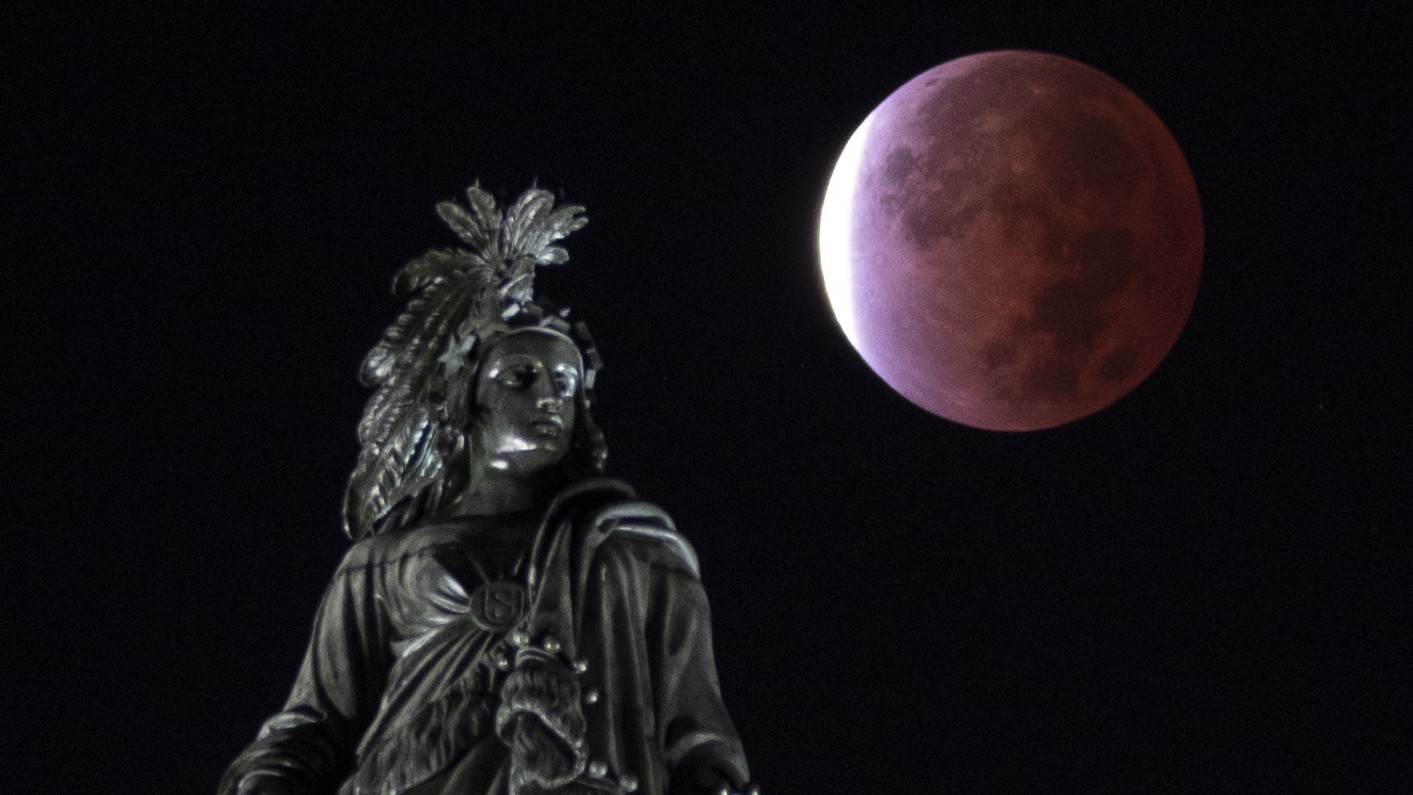 An eclipsed full moon is photographed in a dark sky next to a statue.