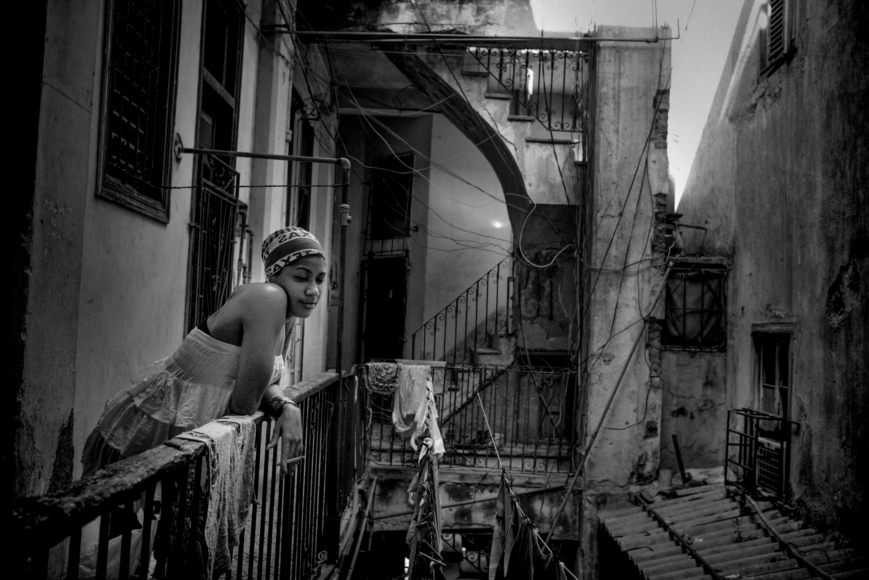 Black and white photograph of a woman leaning over a wrought-iron balcony railing in a Havana alleyway, laundry hanging on the railings around her.
