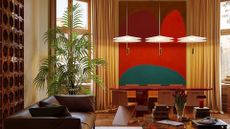 A living room with a brown leather couch on an area rug with a colorful coffee table holding a music ear, a lamp, a vase of flowers, and books. There is a palm plant in the corner of the room near a wood dining set with three pendant lights against a color-blocked wall