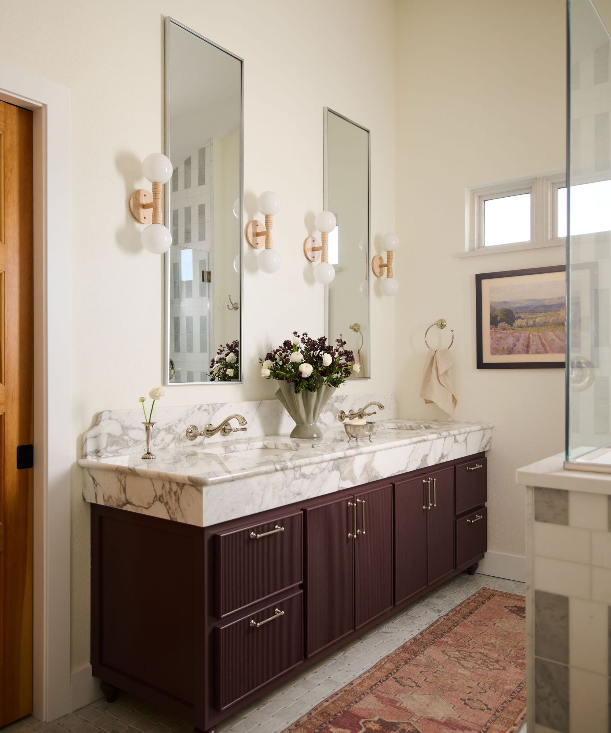 Bathroom with burgundy cabinetry, marble sink, marble floor, pink persian rug and two large mirrors with round sconces