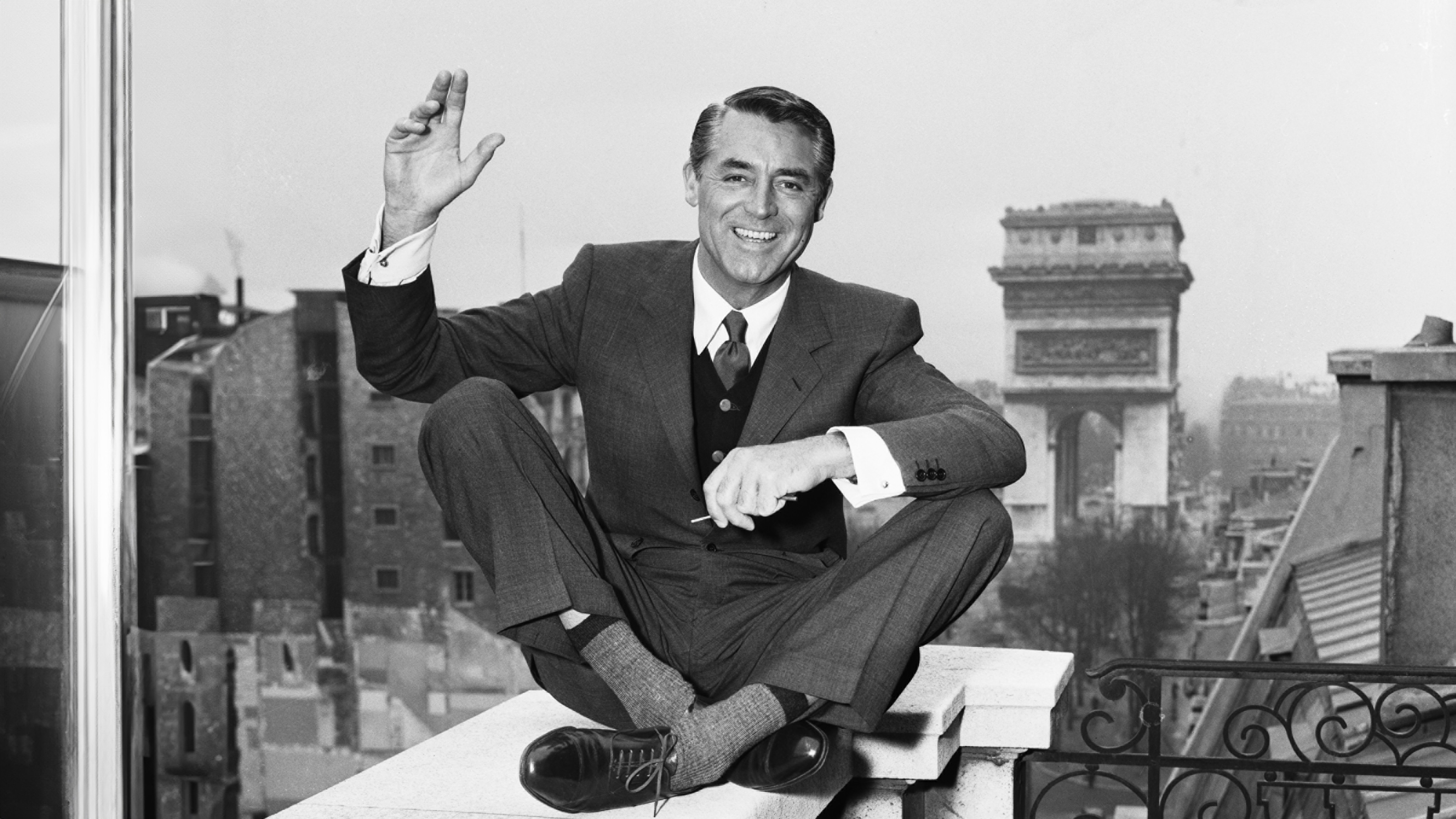 Cary Grant cross legged on a balcony ledge and waving to the camera with his right hand. He is wearing a three-piece suit