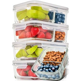 Four glass food containers with divided compartments and glass lids filled with fresh fruit and nuts stacked on top of one another. 