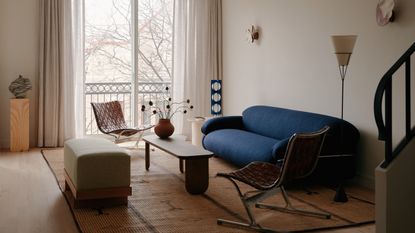 small living room with windows opening out to balcony, plinth in corner, long ottoman, two woven armchair, timber coffee table, and blue sofa 