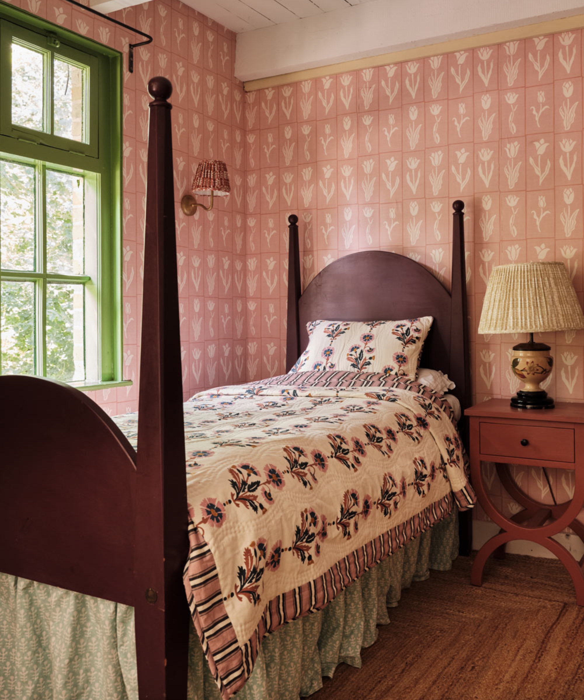 A floral bedroom with pink botanical wallpaper, a dark wood four-poster bed, patterned bedding and a pleated wall lampshade.