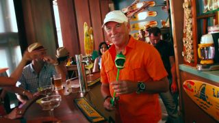 Jimmy Buffett standing behind the bar with a dinosaur toy in hand on the set of Jurassic World.