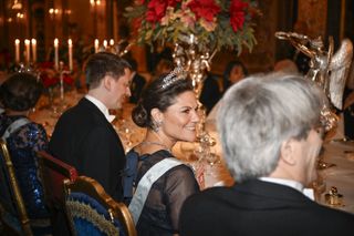 The back of Crown Princess Victoria's head wearing a navy gown and a sash sitting at a candlelit table with flowers