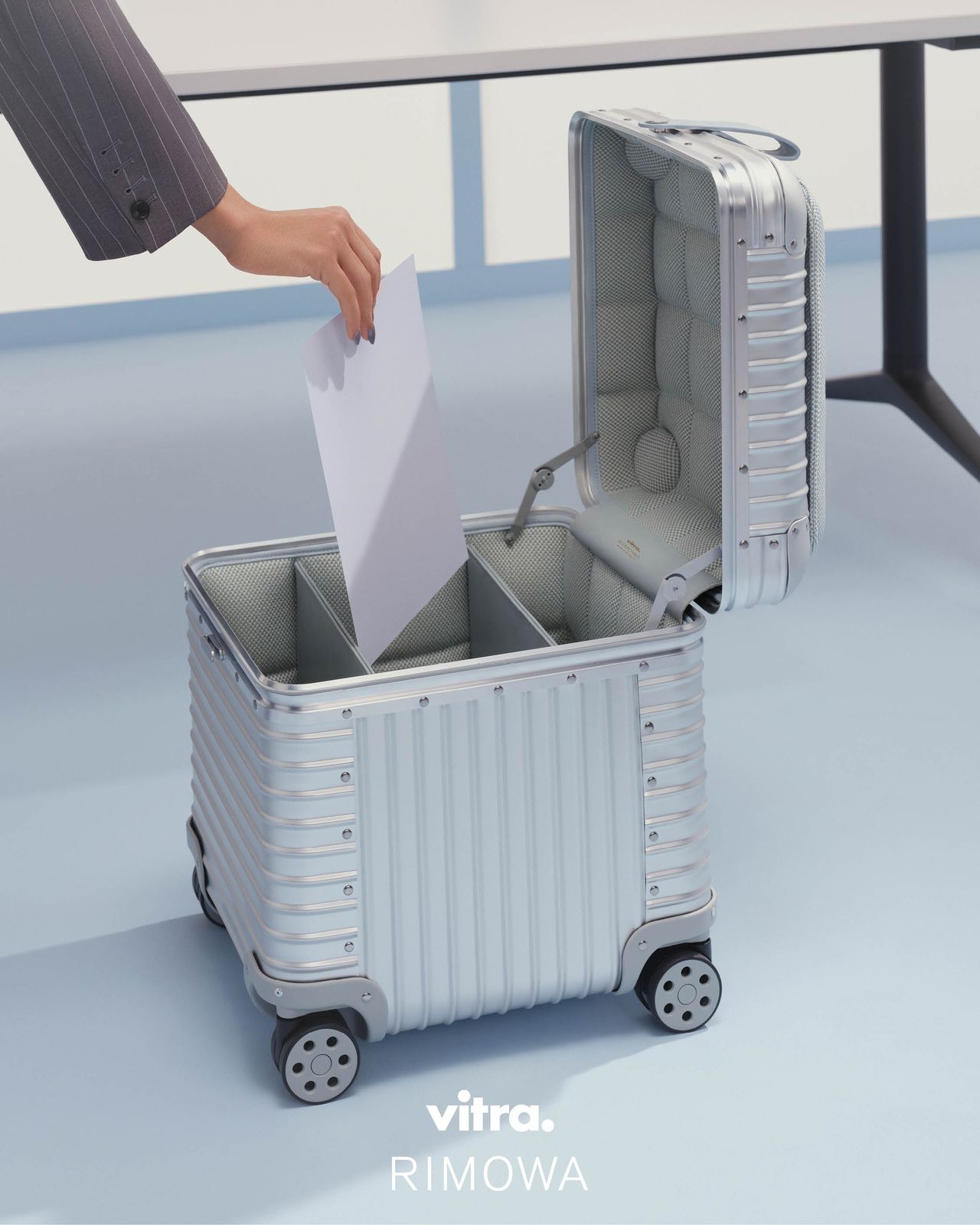 An aluminum cube with wheels has its lid open and a woman is slotting a piece of paper inside