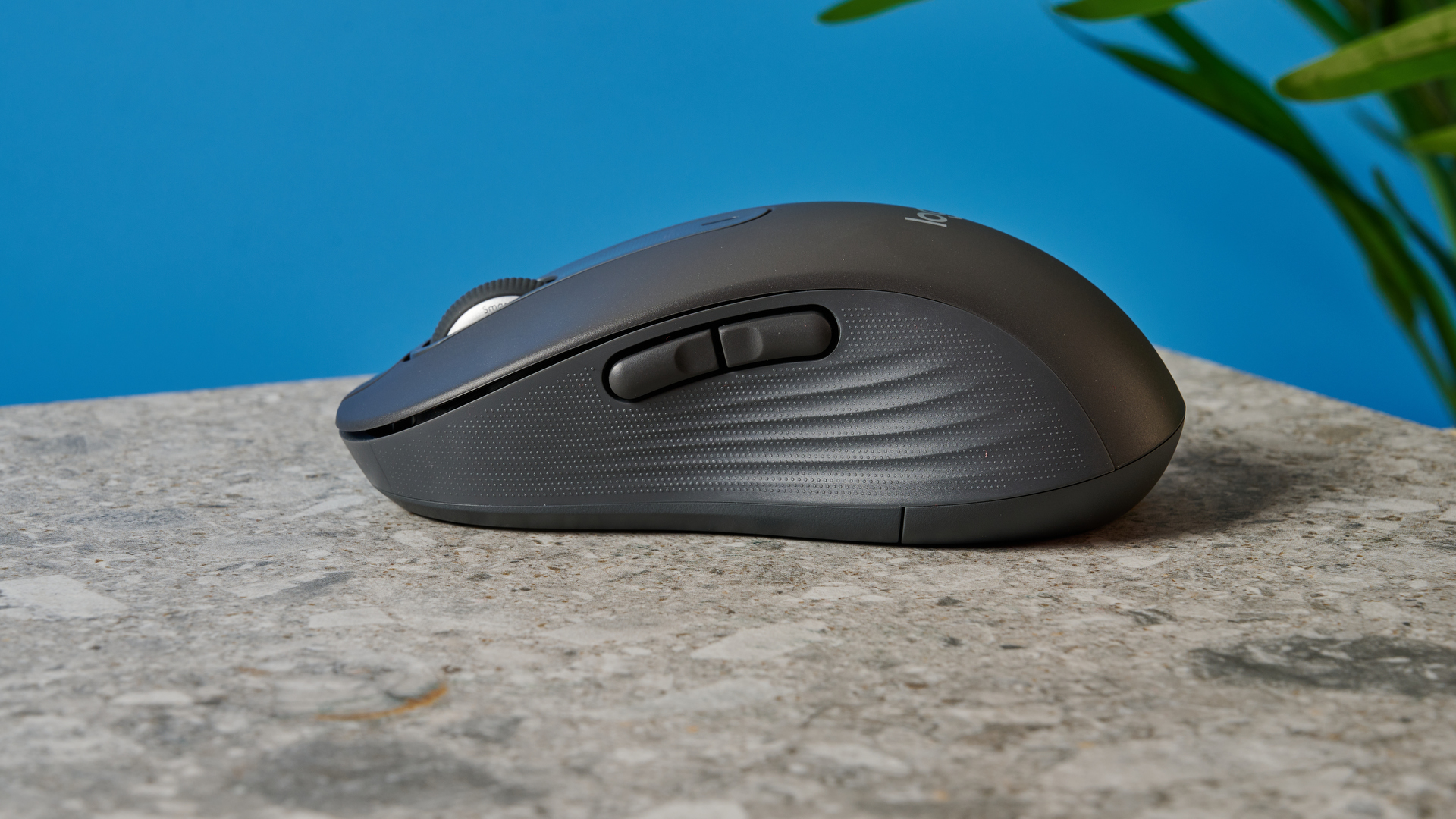 a black mouse with a scroll wheel, two side buttons, and grooved edges, with a logi logo, sits atop a pale table with a blue background