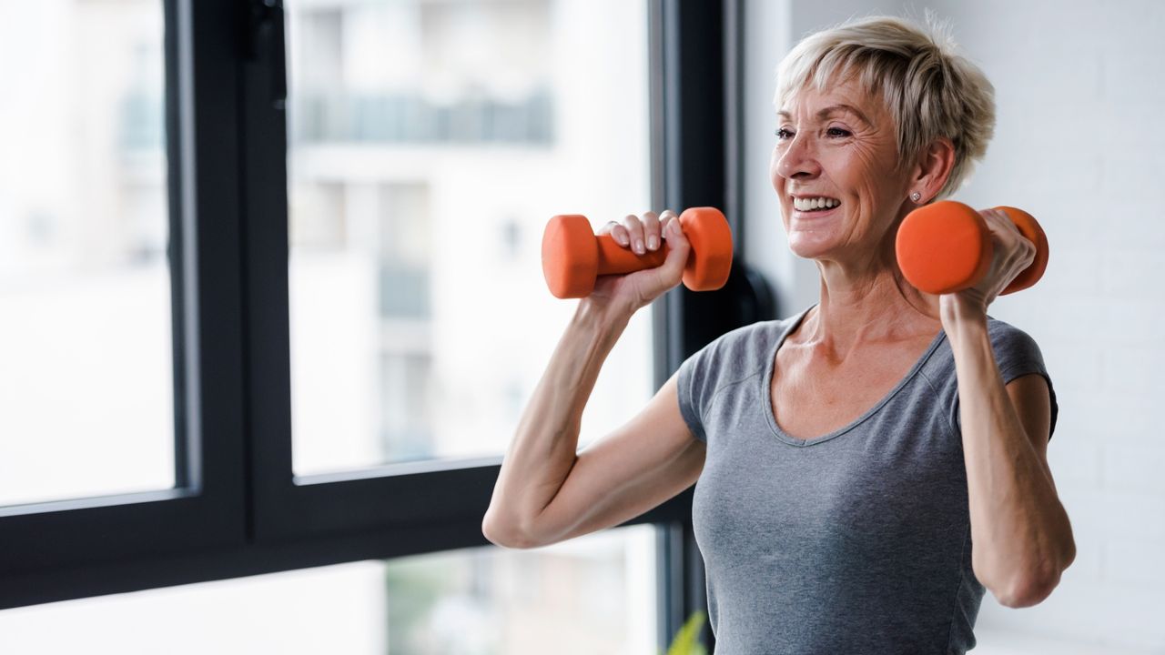 woman in her 50s standing holding two light orange dumbbells over her shoulders and smiling. she&#039;s wearing a grey tshirt and there&#039;s a large window behind her.