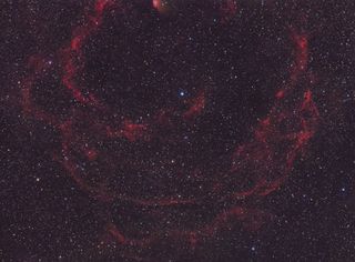 Diffuse threads of red gas and dust reach out into the cosmos against a backdrop of stars.