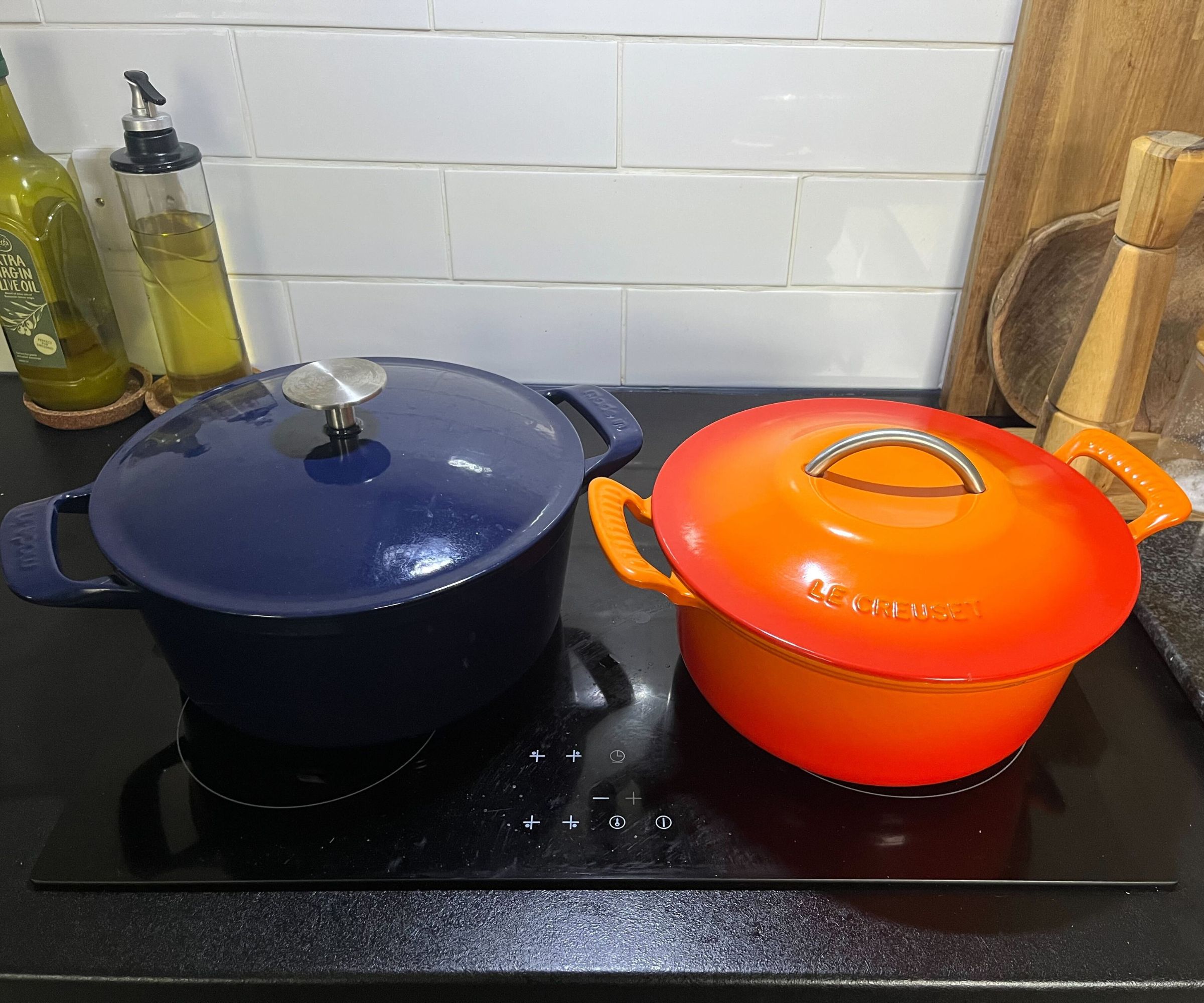 An orange Le Cruset Dutch oven beside a navy blue Made In Dutch oven on a black stovetop