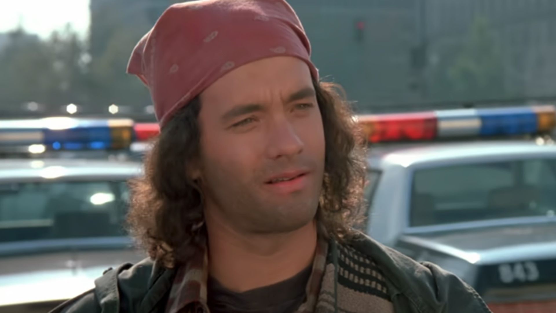 A close up of Tom Hanks with long hair, wearing a red bandana on his head, with a five o'clock shadow with police cars behind him in the opening scene on Dragnet.