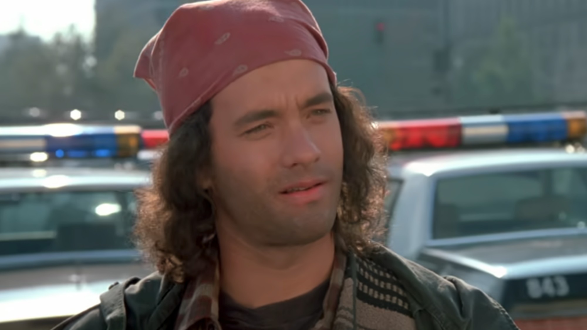 A close up of Tom Hanks with long hair, wearing a red bandana on his head, with a five o'clock shadow with police cars behind him in the opening scene on Dragnet.