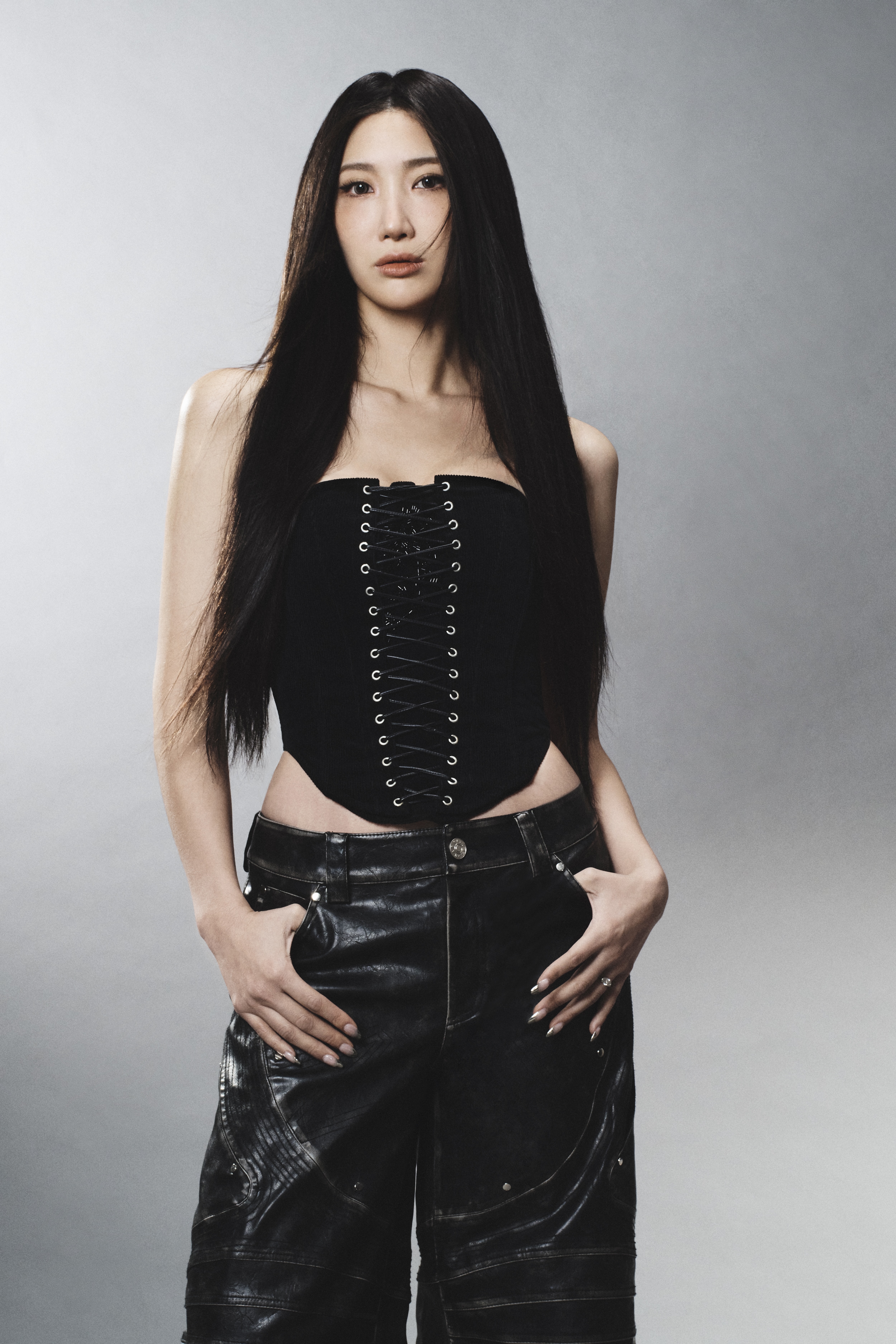 A portrait of singer-songwriter EJAE, wearing a lace-up corset and shiny black pants while standing in front of a gray background.