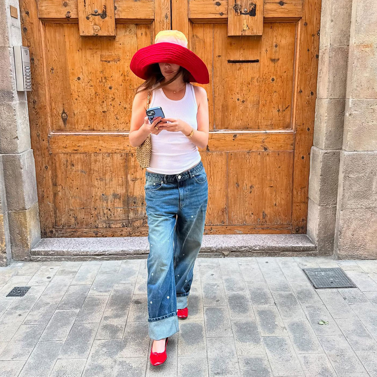 @helenacuesta on phone in summer outfit, big hat, white tank, baggy jeans and red flats