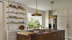 A kitchen with white shaker cabinetry, an island with a red marble countertop, and brass shelving storing kitchenware 