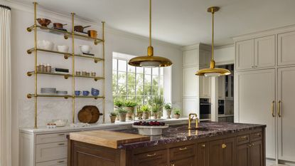A kitchen with white shaker cabinetry, an island with a red marble countertop, and brass shelving storing kitchenware