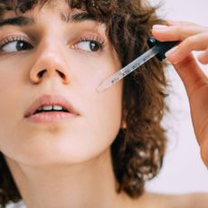 best face serums - woman with shorted curly hair looking off to the side with a pipette of serum next to her face