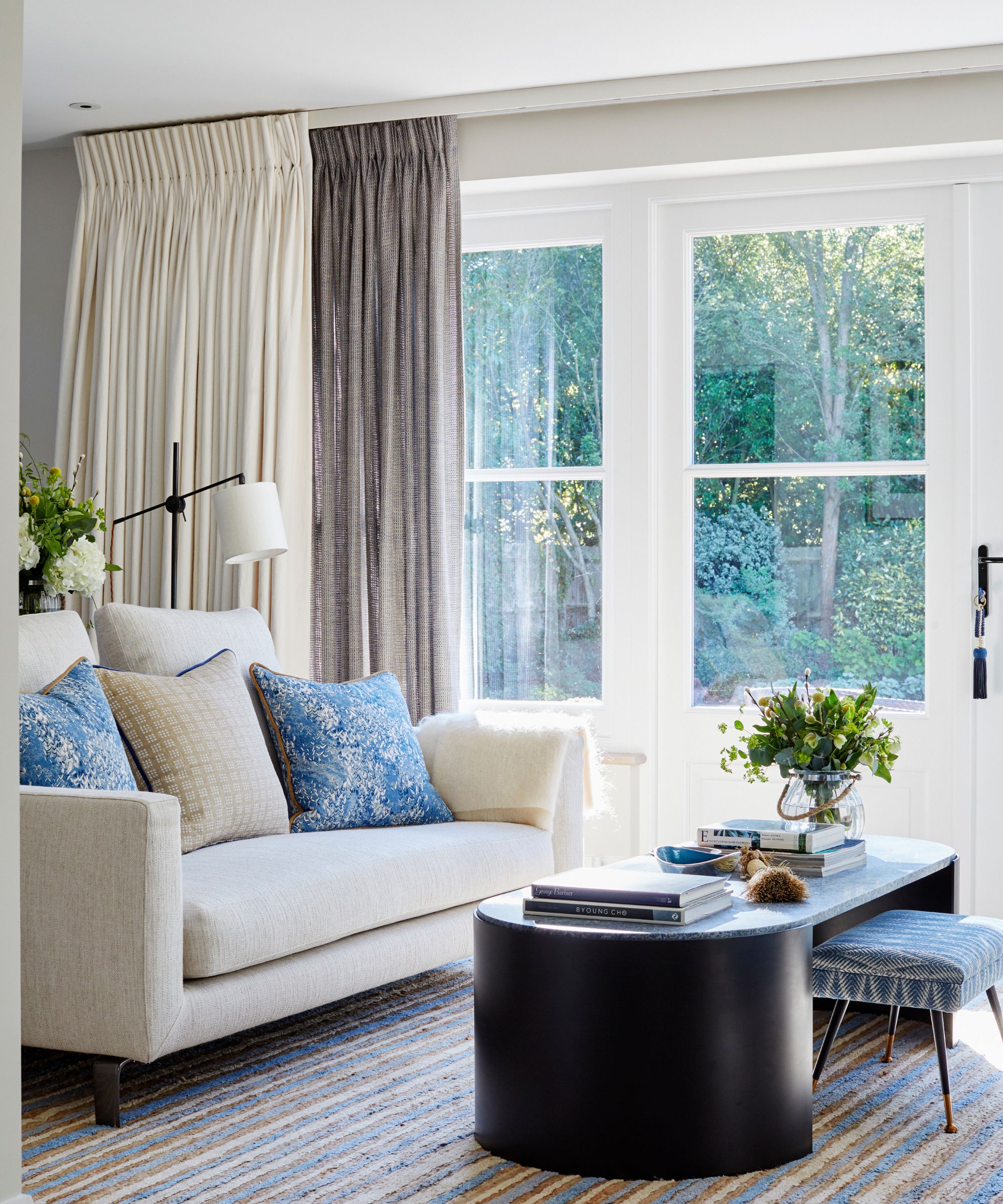 A bright white living room with a white couch with blue cushions, a black coffee table with flowers, and a striped rug. Behind is a large window with curtains.