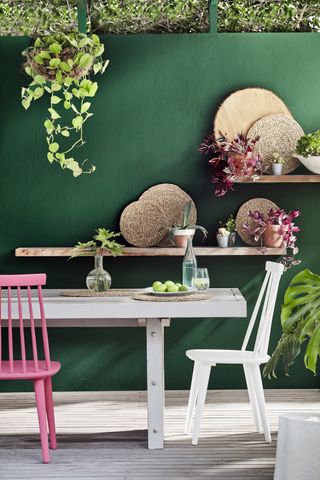 Fence decorating ideas in dark green behind a white table with white and pink painted chairs.