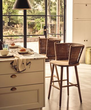 Wicker and wood barstools near marble topped kitchen island, view of garden through crittall doors