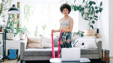 A woman stands in a plant-filled living room in workout clothes, looking at a laptop on a coffee table and holding a resistance band in her hands. The ends of the band is in her hands, while the middle is taught on the floor underneath her foot. Behind her we see a couch and cushions.