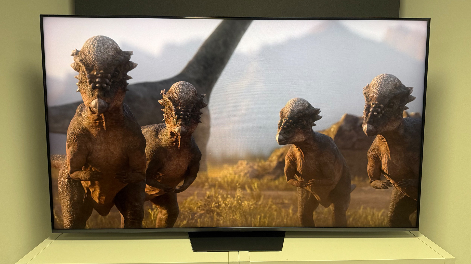 The 65-inch Hisense UR9 RGB Mini LED TV, photographed on a white, wooden unit. On the screen is a still from Netflix documentary, The Dinosaurs.