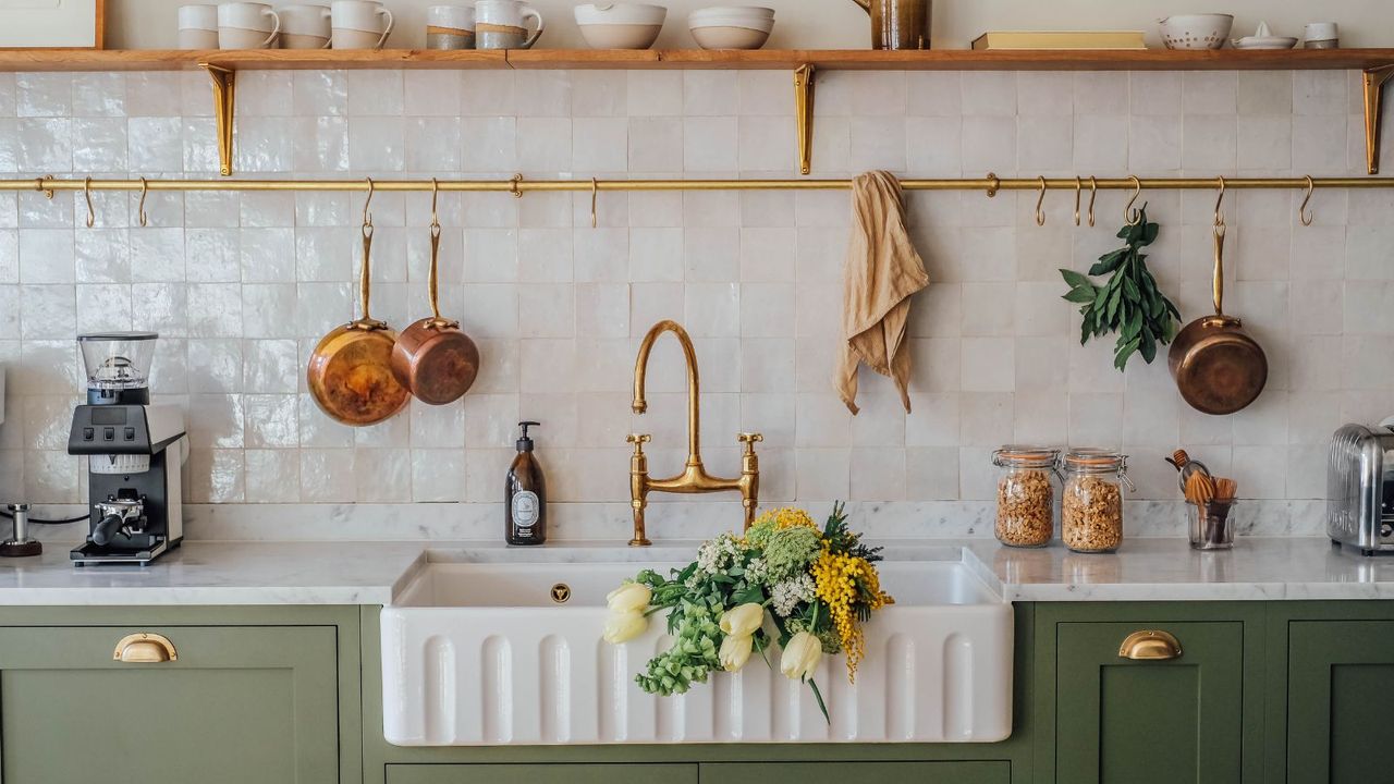 Cream tiled backsplash with china fluted sink, green wooden cupboards and gold rail with copper pans hanging