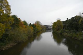 An image showing a river surrounded by trees with a football stadium in the distance