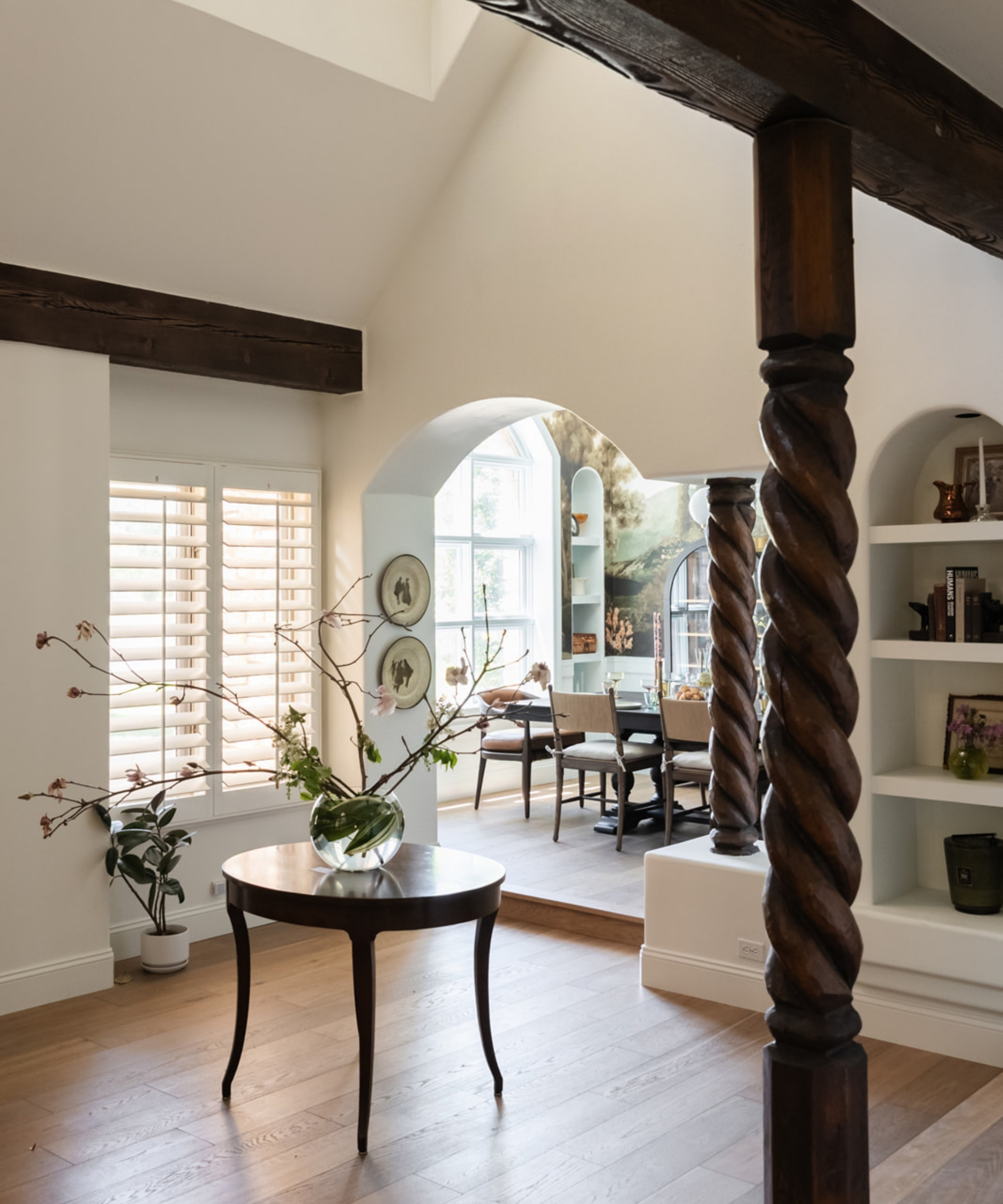 Bright home interior with wooden floors and beams. Pictured in the middle of the room is a small wooden table with a glass-potted plant; a built-in bookshelf is to the right; windows and open shutters are pictured to the left. In the background, a dining room is visible through an archway.