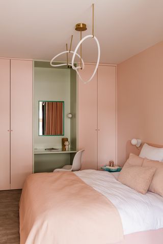 bedroom painted in soft pale pink with vanity table between closets painted in a safe green with a mirror and chair. a modern pendant light hangs over the bed made with pink and white sheets