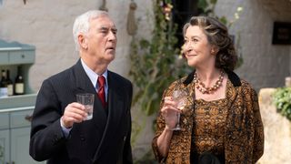 Walter Sullivan (Denis Lawson), wearing a black pinstripe double-breasted suit, stands outside a pub raising a glass of clear liquid in a toast. His wife Grace (Cherie Lunghi) stands next to him, holding a glass of wine, wearing a black and orange patterned two-piece outfit. She is looking at him and smiling.