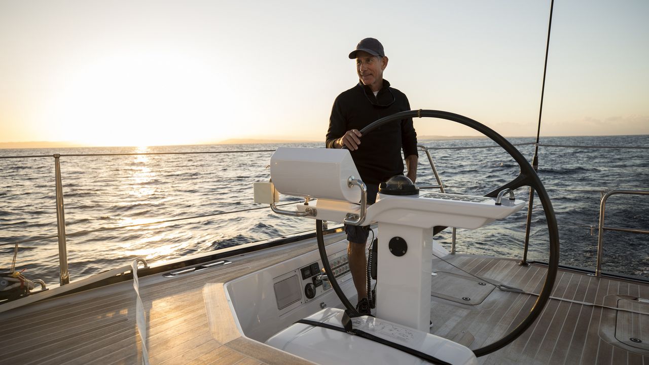 An older man at the helm of a yacht on the open water at sunset.