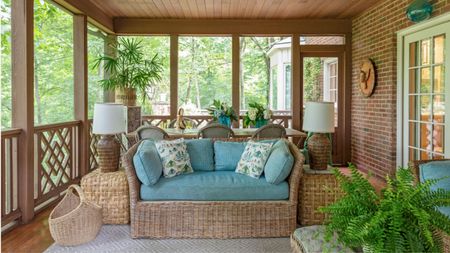 Porch with wooden balcony and ceiling, wicker sofa with blue cushions, wicker side tables with lamps, ferns, and a dining table with cane dining chairs behind.