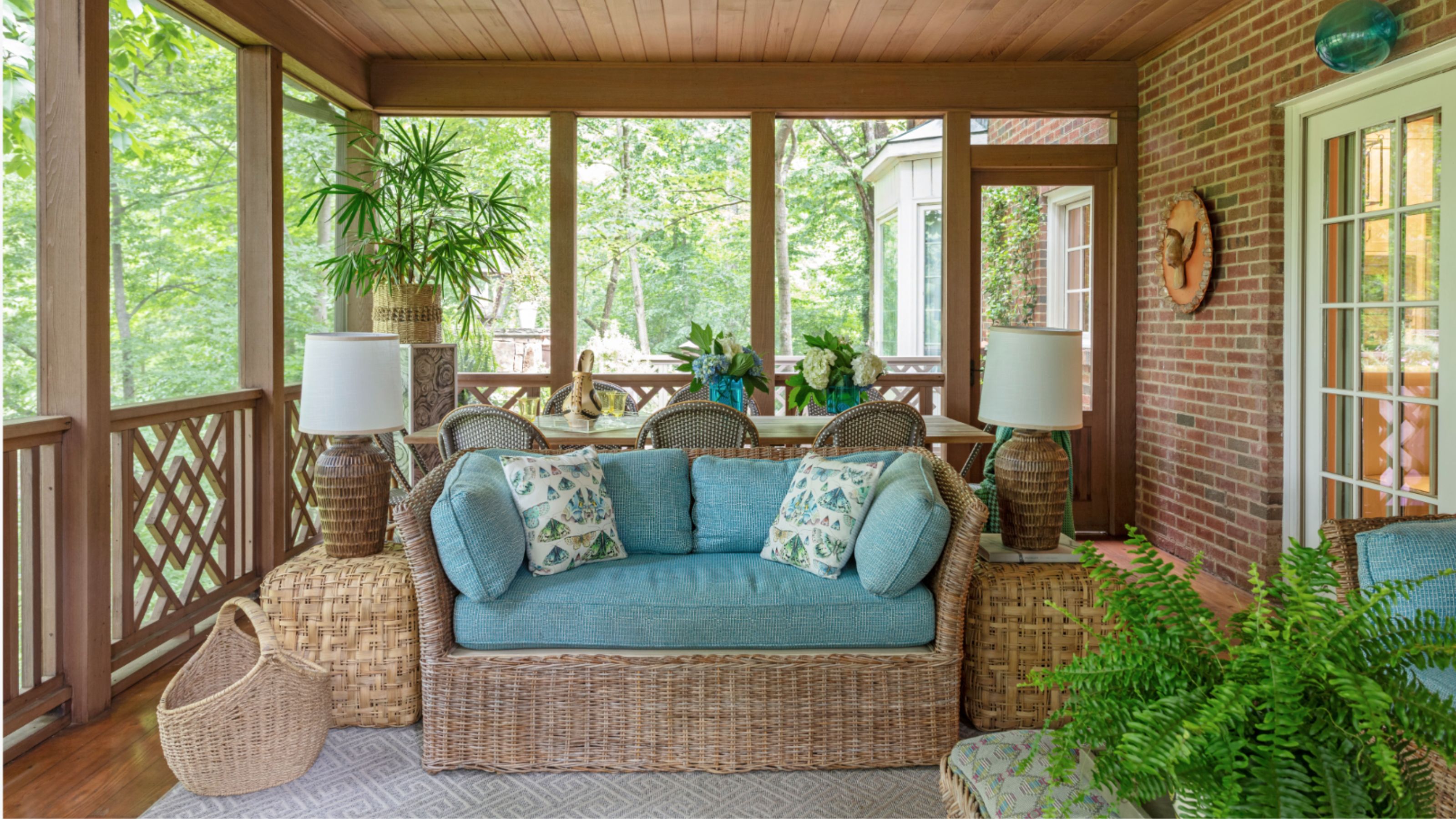 Porch with wooden balcony and ceiling, wicker sofa with blue cushions, wicker side tables with lamps, ferns, and a dining table with cane dining chairs behind.