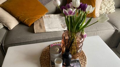 Annie's white coffee table with round seagrass matt in the middle and Real Homes magazine on top, with a black reed diffuser and two white candles beside a vase filled with colorful tulips
