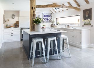 kitchen with old beams and white cabinets with black island and silver bar stools and grey tiled floor old master and advertising signon the white wall