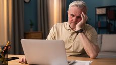 A tired and frustrated-looking older man sits in front of his laptop at the dining room table.