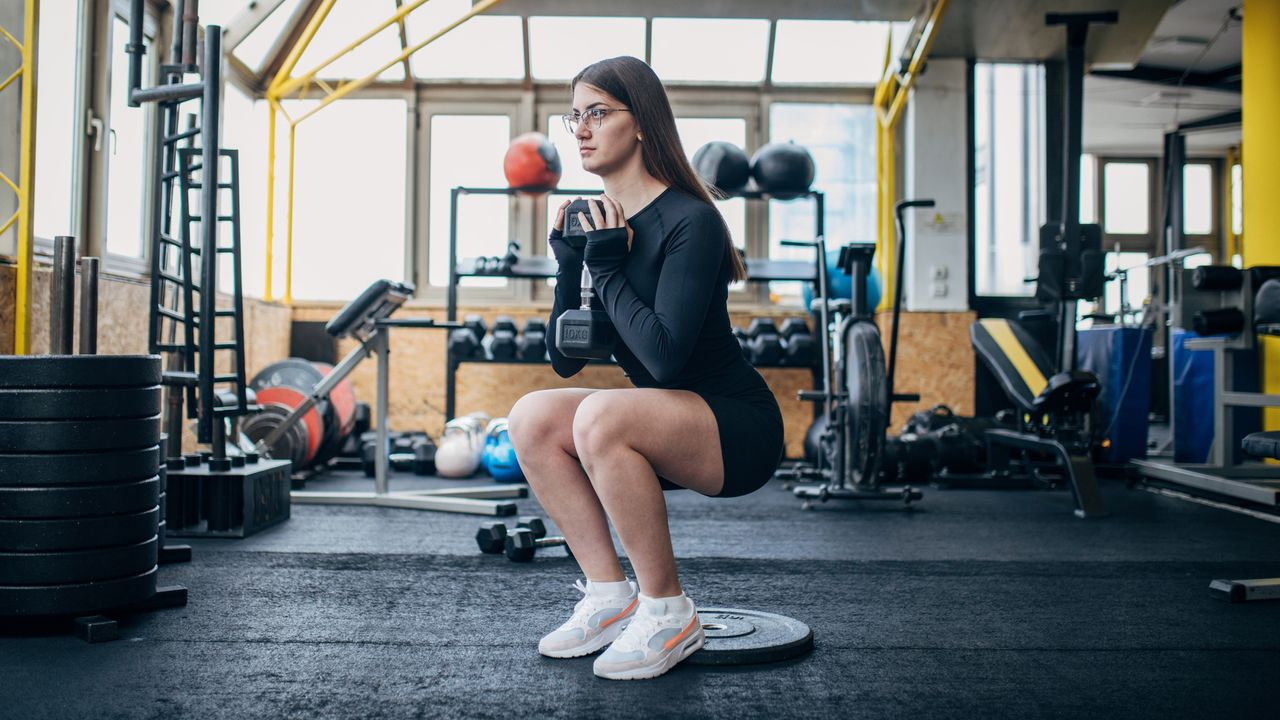 Woman performing squat in a gym with her heels raised on a weight plate, holding one end of a dumbbell in front of her chest