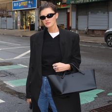 style influencer Coco Schiffer wearing minimalist outfit idea with a white T-shirt, black coat, black v-neck sweater, jeans, and The Row top handle black tote bag