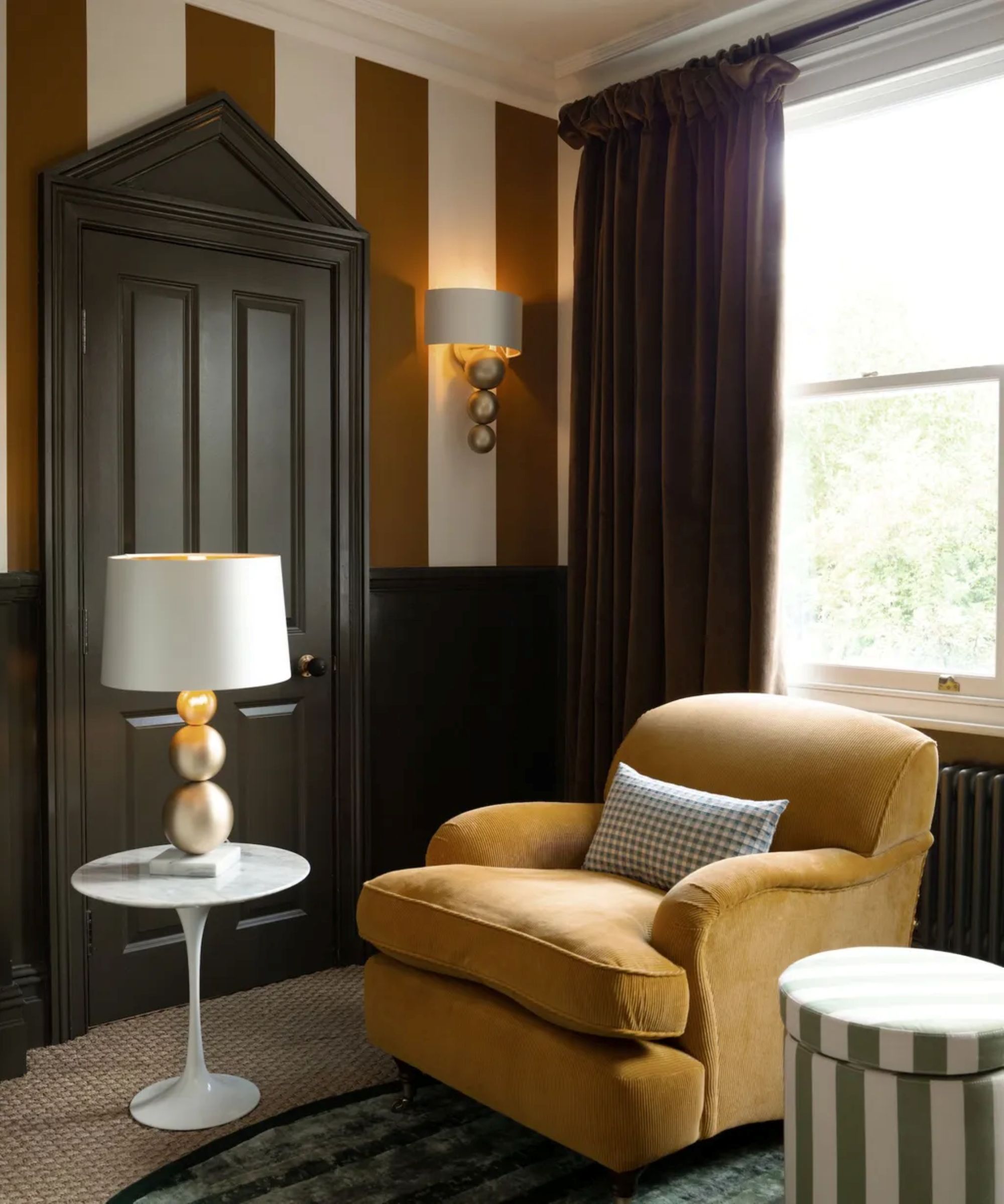 a reading corner with a mustard yellow chair, a striped ottoman, and small side table with a table lamp in a room with striped brown and white walls