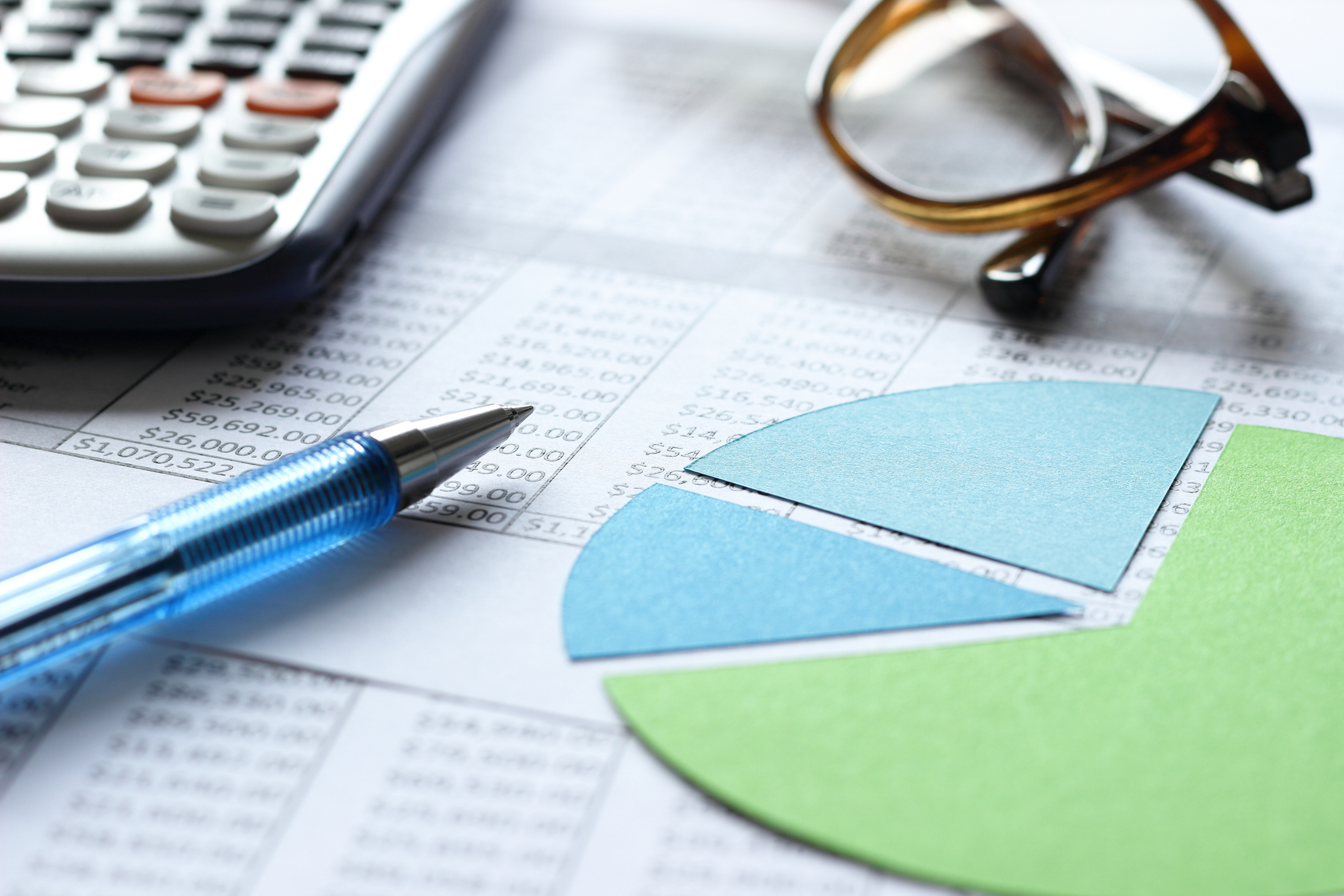 green and blue pie chart on top of spreadsheets and next to a calculator, pen and pair of eyeglasses